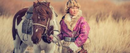 A nomad woman in a Mongolian grassland field with her horse. A Mongolian woman holding her horse in a field as the wind blows against her face and the tall grass